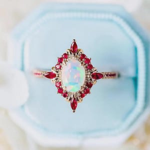 Peut inclure: Bague en or ornée d'une pierre centrale ovale en opale, entourée de petites pierres rouges disposées en forme de losange. La monture de la bague est également ornée de petites pierres rouges. Présentée dans un écrin bleu clair.