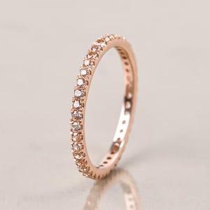 May include: Rose gold eternity ring with a continuous row of small, clear gemstones. The ring's band is a warm, metallic color, and the gemstones sparkle. The ring is set against a soft, neutral background.