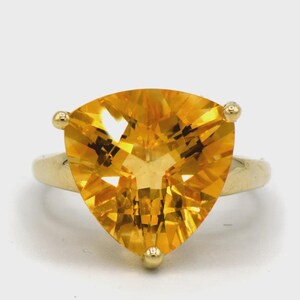 May include: A gold ring featuring a large, triangular, faceted, yellow gemstone. The gemstone is the focal point, with a brilliant, radiant appearance. The ring's band is a simple, polished gold, and the gemstone is secured with three small gold prongs.