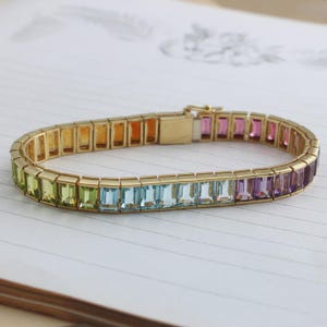 Puede incluir: Una pulsera dorada con un espectro de piedras preciosas de colores. Las piedras de corte rectangular transitan del verde al azul, luego al rosa y al morado. La pulsera está engarzada en una banda dorada con un cierre seguro.