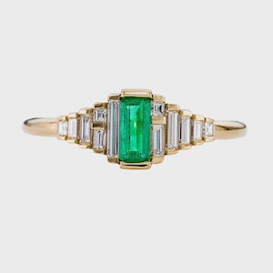 Può includere: Un anello d'oro con uno smeraldo rettangolare centrale e diamanti taglio baguette. L'anello ha un design geometrico. Lo smeraldo è di un verde vibrante e i diamanti sono incastonati in una montatura d'oro.