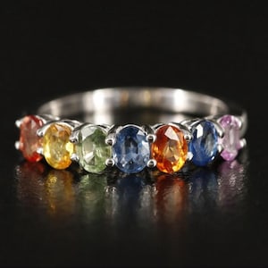 Puede incluir: Un anillo de plata con una fila de piedras preciosas ovaladas en colores del arcoíris: rojo, amarillo, verde, azul, naranja y morado. Las piedras están engastadas en garras individuales, creando un diseño vibrante. Fondo negro.