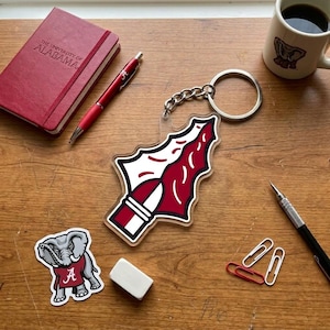 May include: A clear acrylic keychain featuring a red and white design, with a silver chain and ring. The keychain is next to a red notebook with the text "THE UNIVERSITY OF ALABAMA", a red pen, and a sticker of an elephant wearing a red shirt.