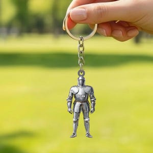 May include: A silver-colored knight keychain with the name "BRIAN" on the chest. The knight is wearing full armor and is attached to a silver keyring and chain. The background is a blurred green.
