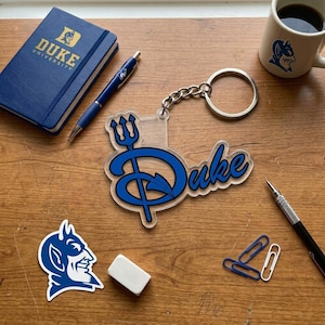 May include: An assortment of Duke University merchandise, including a blue notebook, pen, keychain, coffee mug, and stickers. The keychain features the word "Duke" in a stylized font with a trident and devil graphic.