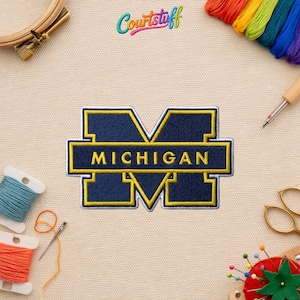 May include: Embroidered patch featuring the word "MICHIGAN" in yellow lettering, set against a navy blue background. The patch has a yellow border and a stylized "M" design. The patch is surrounded by sewing supplies, including thread spools and a needle.