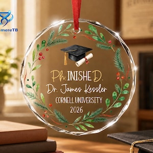 May include: A circular clear glass ornament featuring a printed wreath of green pine branches and red berries. In the center, a black graduation cap and a rolled diploma are depicted above the text Ph. INISHED. Dr. James Kessler CORNELL UNIVERSITY 2026.