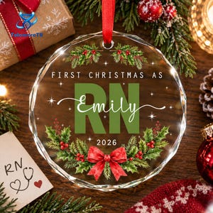 May include: A circular, faceted glass Christmas ornament featuring a red ribbon hanger. The design includes green holly leaves, red berries, and a red bow. Text reads: FIRST CHRISTMAS AS RN, Emily, and 2026, accented with small white stars.
