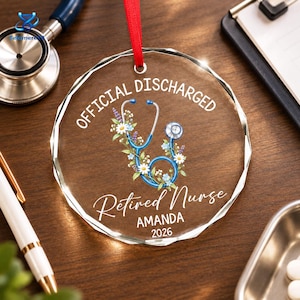May include: A circular clear glass ornament with a red ribbon hanger. The text reads OFFICIAL DISCHARGED, Retired Nurse, AMANDA, and 2026. A blue stethoscope is illustrated in the center, intertwined with white and blue flowers and green leaves.