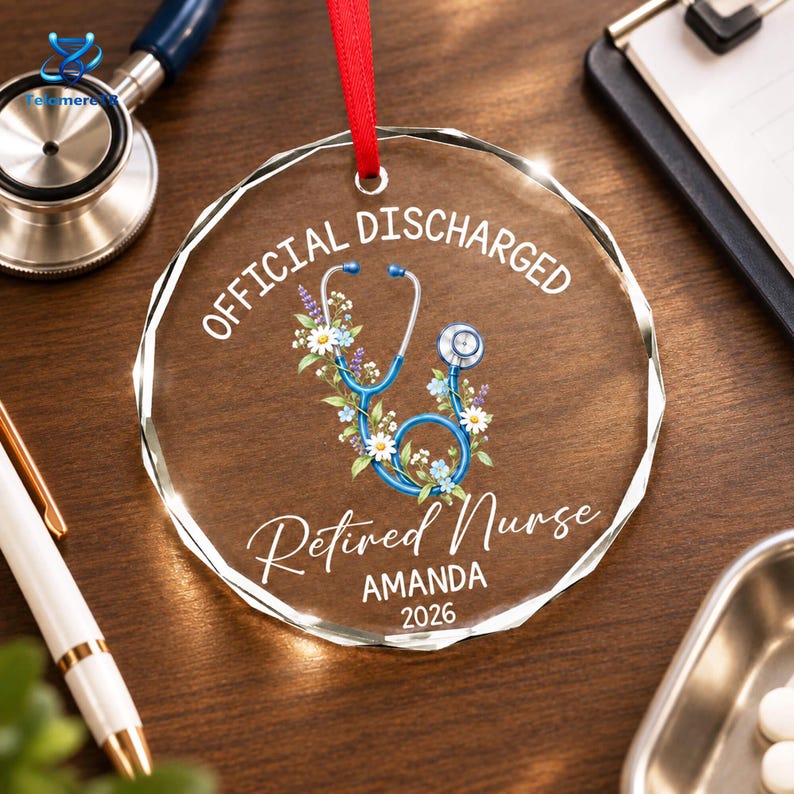 May include: A circular clear glass ornament with a red ribbon hanger. The text reads OFFICIAL DISCHARGED, Retired Nurse, AMANDA, and 2026. A blue stethoscope is illustrated in the center, intertwined with white and blue flowers and green leaves.