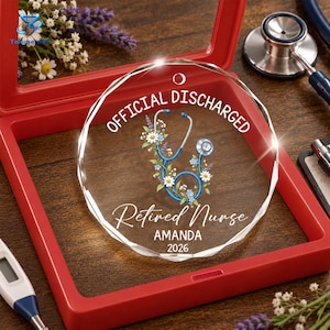 May include: A circular clear acrylic ornament featuring a blue stethoscope intertwined with white daisies and purple lavender. The text reads OFFICIAL DISCHARGED, Retired Nurse, AMANDA, and 2026. It has a small hole at the top for hanging.