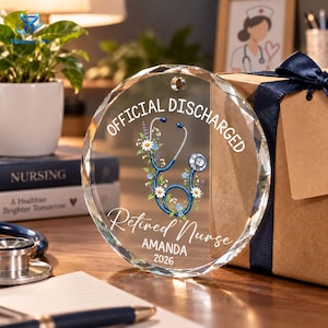 May include: A circular clear glass ornament featuring a blue stethoscope graphic surrounded by white and blue flowers. The text reads OFFICIAL DISCHARGED, Retired Nurse, AMANDA, and 2026. The ornament has a small hole at the top for hanging.