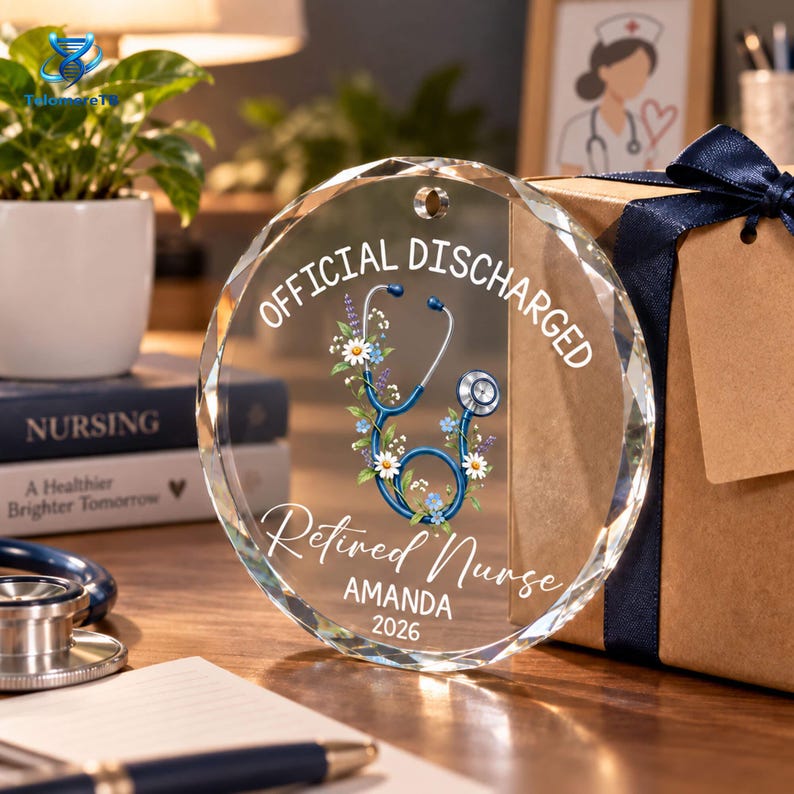 May include: A circular clear glass ornament featuring a blue stethoscope graphic surrounded by white and blue flowers. The text reads OFFICIAL DISCHARGED, Retired Nurse, AMANDA, and 2026. The ornament has a small hole at the top for hanging.