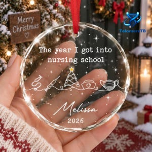May include: A clear, circular, faceted glass Christmas ornament hanging from a red ribbon. It features the white text 'The year I got into nursing school', a line drawing of a stethoscope, syringe, tree, nurse cap, mask, the name 'Melissa', and '2026'.