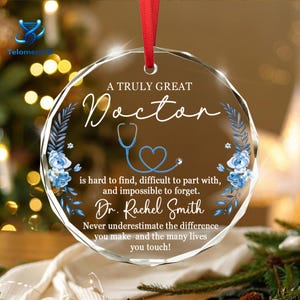 May include: A circular, faceted glass ornament hanging from a red ribbon. It features blue floral illustrations, a blue stethoscope shaped like a heart, and the text: A TRULY GREAT Doctor is hard to find, difficult to part with, and impossible to forget. Dr. Rachel Smith. Never underestimate the difference you make and the many lives you touch!