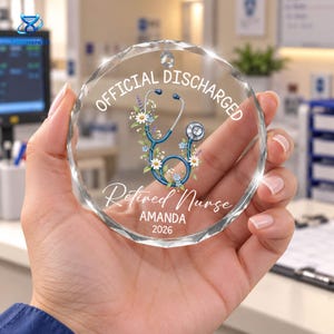 May include: A circular clear glass ornament with faceted edges featuring a blue stethoscope graphic intertwined with white and purple flowers. The text reads OFFICIAL DISCHARGED, Retired Nurse, AMANDA, and 2026 in white lettering on the surface.