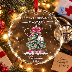 May include: A round, clear, faceted glass ornament featuring a printed graphic of a Christmas tree decorated with a pink stethoscope, a nurse cap, and a pink bow. The text reads THE YEAR I BECAME A nurse, Amanda, and 2026, with snowflakes.