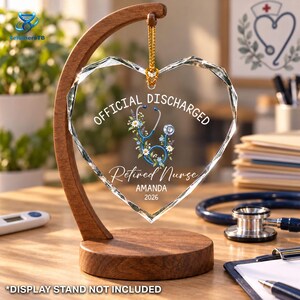May include: A heart-shaped clear acrylic ornament hanging from a curved wooden stand. The ornament features a blue stethoscope graphic with flowers, the text "OFFICIAL DISCHARGED", "Retired Nurse", "AMANDA", and "2026" printed in white.