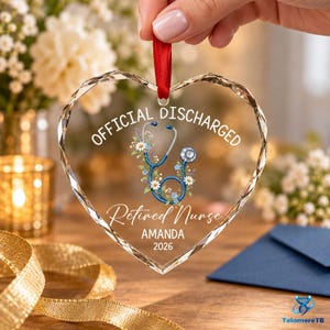 May include: A heart-shaped clear acrylic ornament with a red ribbon hanger. The text reads OFFICIAL DISCHARGED, Retired Nurse, AMANDA, and 2026. A blue stethoscope graphic is intertwined with white and purple flowers in the center of the ornament.