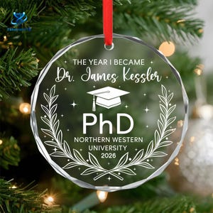 May include: A clear, circular, faceted glass ornament featuring white etched text that reads THE YEAR I BECAME Dr. James Kessler, PhD, NORTHERN WESTERN UNIVERSITY 2026, with a graduation cap icon and laurel branches, suspended by a red ribbon.