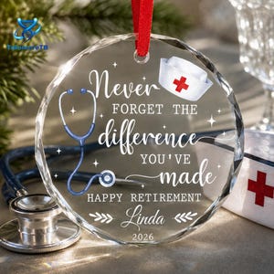 May include: A circular clear glass ornament with a red ribbon hanger featuring a white nurse cap, a blue stethoscope, and the text: Never FORGET THE difference YOU'VE made HAPPY RETIREMENT Linda 2026. The design includes small star accents.