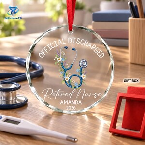 May include: A circular clear glass ornament with a red ribbon hanger, featuring a blue stethoscope graphic intertwined with colorful wildflowers. The text reads OFFICIAL DISCHARGED, Retired Nurse, AMANDA, and 2026 in white lettering.