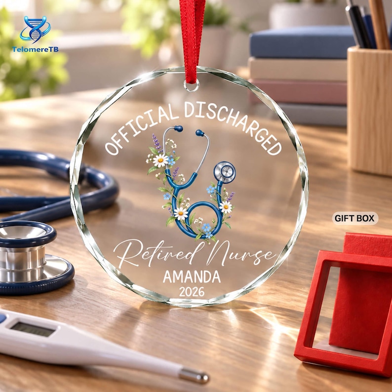 May include: A circular clear glass ornament with a red ribbon hanger, featuring a blue stethoscope graphic intertwined with colorful wildflowers. The text reads OFFICIAL DISCHARGED, Retired Nurse, AMANDA, and 2026 in white lettering.