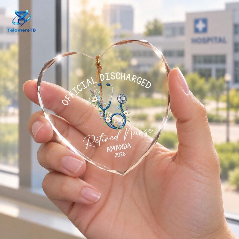 May include: A clear, heart-shaped acrylic ornament featuring a printed blue stethoscope intertwined with white and purple flowers. The text reads OFFICIAL DISCHARGED, Retired Nurse, AMANDA, and 2026 in white lettering, suspended by a gold cord.