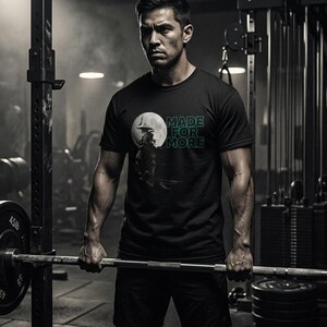 以下が含まれることがあります： ジムで、黒いTシャツを着た人がいます。Tシャツには緑色の文字で「MADE FOR MORE」と書かれています。月の前にいる人物のグラフィックが特徴です。その人はバーベルを持っています。