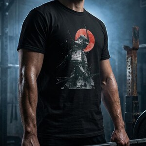 以下が含まれることがあります： 黒いTシャツ。背景に赤い太陽と侍のグラフィック。デザインはグレーと黒の色合いで、印象的なコントラストを生み出しています。柔らかい素材で作られたシャツです。