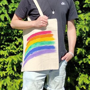 Op de afbeelding: Een beige draagtas met een regenboogontwerp in rood, oranje, geel, groen, blauw en paars. De tas wordt vastgehouden door een persoon die een zwart t-shirt en lichtblauwe shorts draagt. De achtergrond is groen.