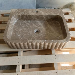 May include: A rectangular, brown marble sink with a scalloped edge. The sink has a central drain hole and a faucet hole. The marble has a natural veining pattern. The sink is displayed on a wooden pallet.