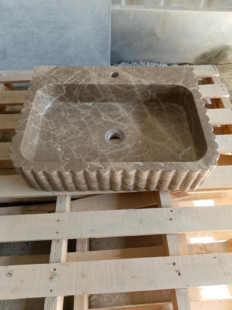 May include: A rectangular, brown marble sink with a scalloped edge. The sink has a central drain hole and a faucet hole. The marble has a natural veining pattern. The sink is displayed on a wooden pallet.