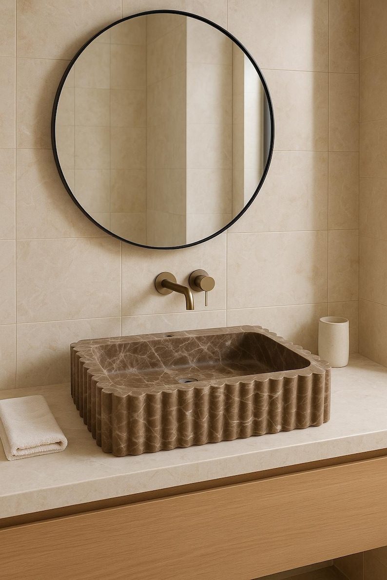 May include: A brown marble bathroom sink with a fluted edge sits on a light-colored countertop. A round black mirror hangs above the sink, with a gold faucet and a small white towel to the left. A white ceramic cup is on the right.