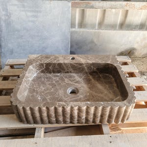 May include: A rectangular, brown marble sink with a scalloped edge. The sink has a central drain hole and a faucet hole. The marble has a natural veining pattern. The sink is on a wooden pallet.