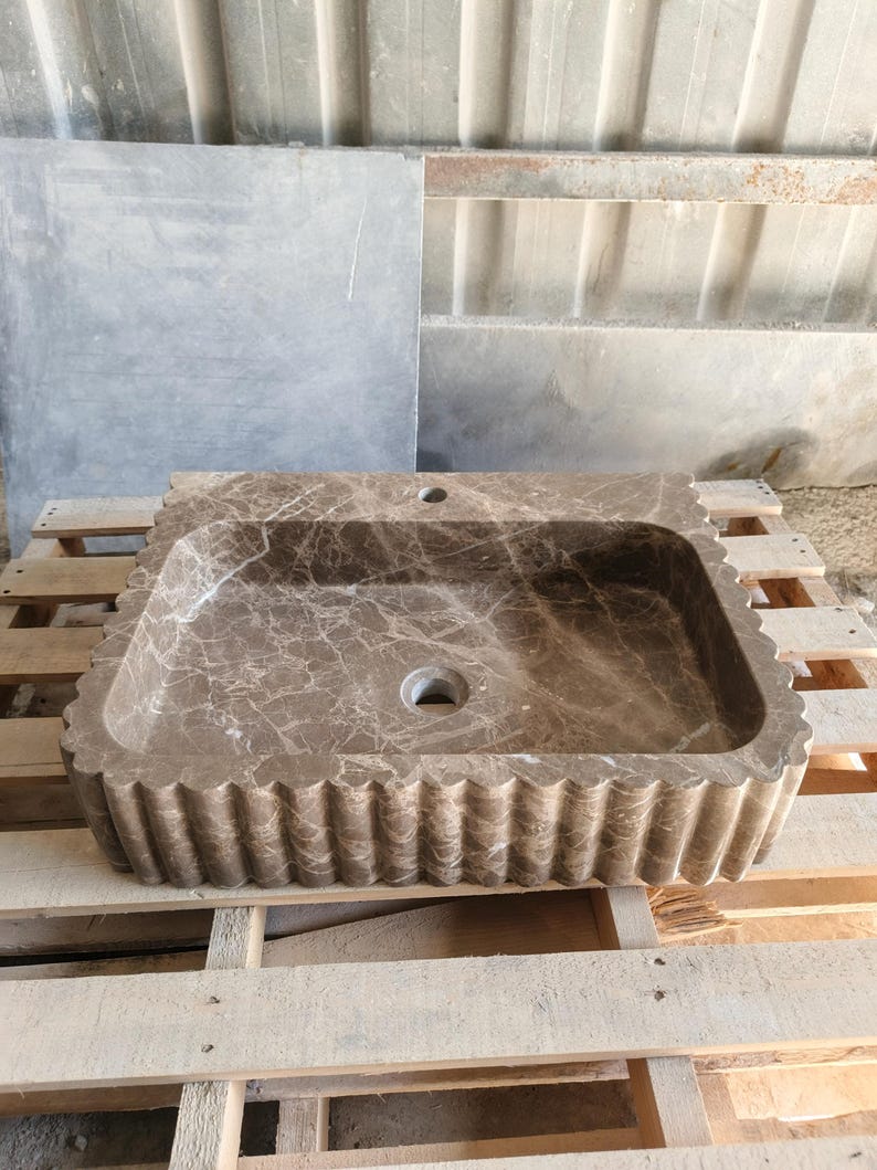 May include: A rectangular, brown marble sink with a scalloped edge. The sink has a central drain hole and a faucet hole. The marble has a natural veining pattern. The sink is on a wooden pallet.