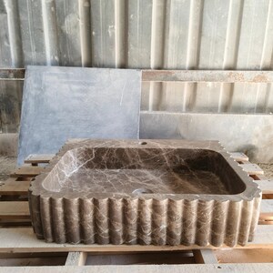May include: A rectangular brown marble sink with a scalloped edge. The sink has a natural stone pattern and is set on a wooden pallet. The sink has a single drain hole.