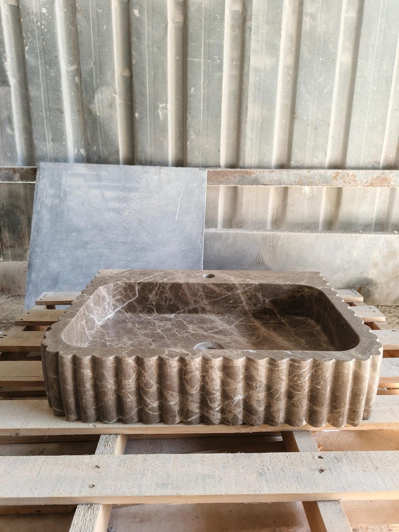 May include: A rectangular brown marble sink with a scalloped edge. The sink has a natural stone pattern and is set on a wooden pallet. The sink has a single drain hole.