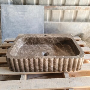 May include: A rectangular brown marble sink with a scalloped edge. The sink has a central drain hole and a faucet hole. The marble has a natural veining pattern. The sink is displayed on a wooden pallet.