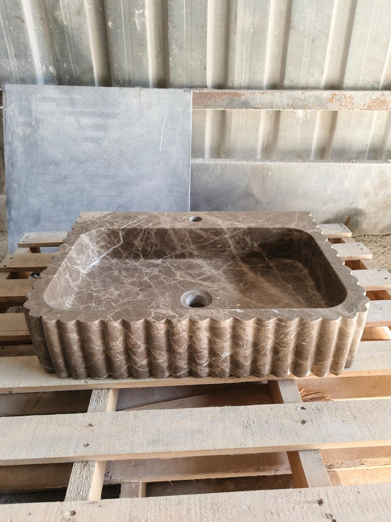May include: A rectangular brown marble sink with a scalloped edge. The sink has a central drain hole and a faucet hole. The marble has a natural veining pattern. The sink is displayed on a wooden pallet.