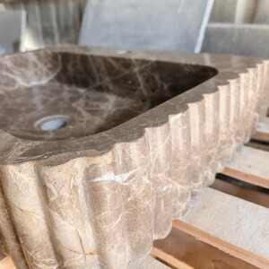 May include: A rectangular, brown marble sink with a scalloped edge. The sink has a central drain hole and a natural veining pattern throughout. The sink is made of stone and is designed for bathroom use.