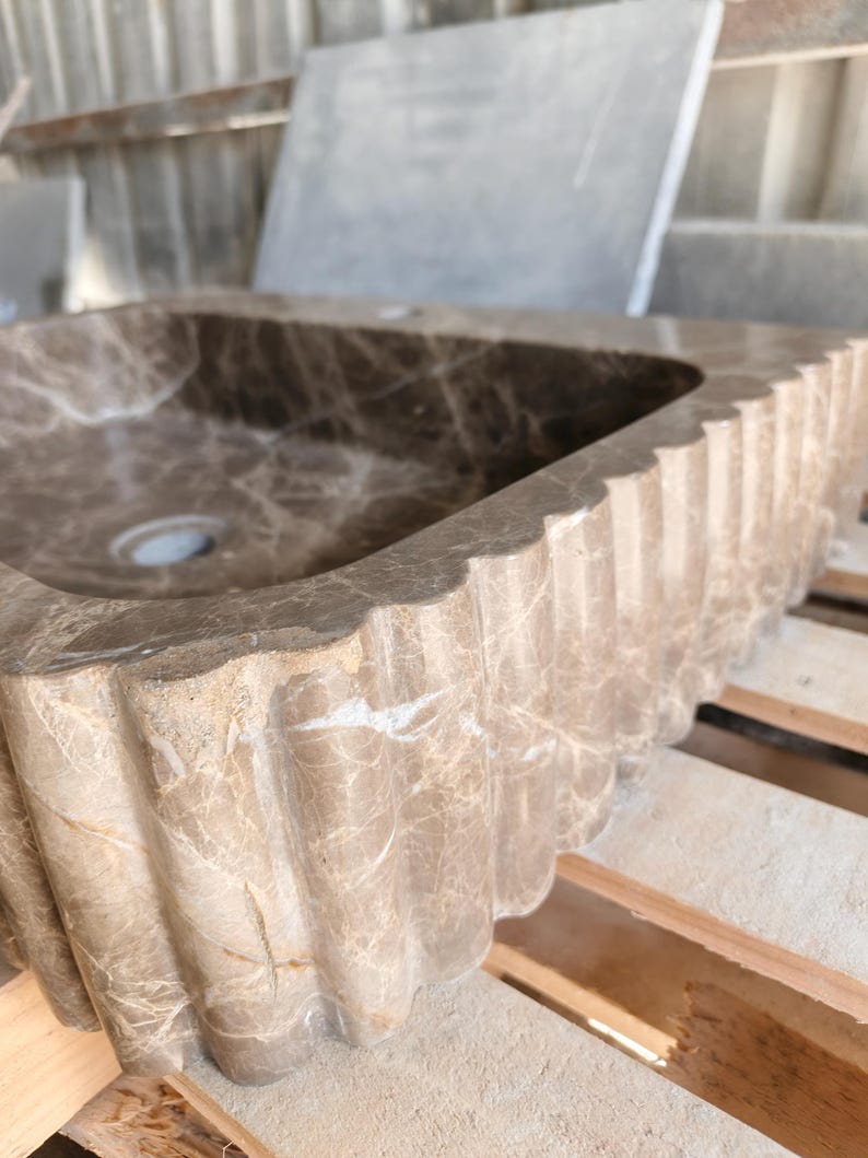 May include: A rectangular, brown marble sink with a scalloped edge. The sink has a central drain hole and a natural veining pattern throughout. The sink is made of stone and is designed for bathroom use.