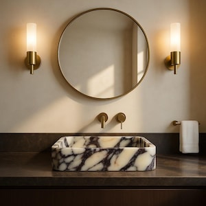 May include: A contemporary bathroom scene with a rectangular marble sink with dark veining, a circular gold-framed mirror, and two wall lights with frosted glass shades. Gold fixtures and a towel rail add to the stylish design.