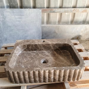 May include: A rectangular, brown marble sink with a scalloped edge. The sink has a central drain hole and a smaller hole for a faucet. The marble has a natural veining pattern, and the sink is set on a wooden pallet.