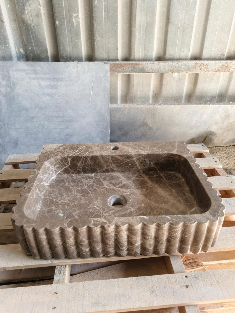 May include: A rectangular, brown marble sink with a scalloped edge. The sink has a central drain hole and a smaller hole for a faucet. The marble has a natural veining pattern, and the sink is set on a wooden pallet.