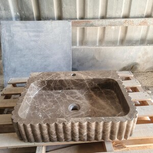 May include: A rectangular, brown marble sink with a scalloped edge. The sink has a natural veining pattern and two drain holes. It is displayed on a wooden pallet.