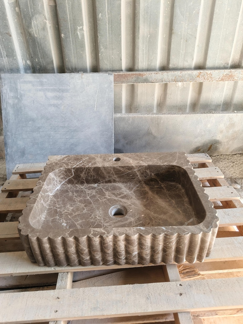 May include: A rectangular, brown marble sink with a scalloped edge. The sink has a natural veining pattern and two drain holes. It is displayed on a wooden pallet.