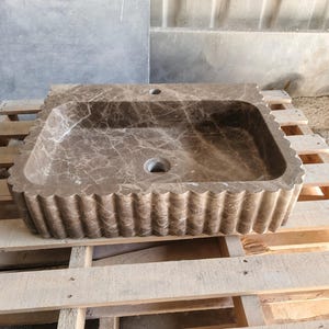May include: A rectangular, brown marble sink with a scalloped edge. The sink has a central drain and a faucet hole. The marble has a natural veining pattern. The sink is displayed on a wooden pallet.