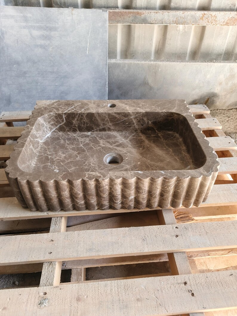 May include: A rectangular, brown marble sink with a scalloped edge. The sink has a central drain and a faucet hole. The marble has a natural veining pattern. The sink is displayed on a wooden pallet.