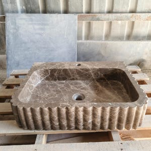 May include: A rectangular brown marble sink with a scalloped edge. The sink has a central drain and a faucet hole. The marble has a natural veining pattern. The sink is displayed on a wooden pallet.
